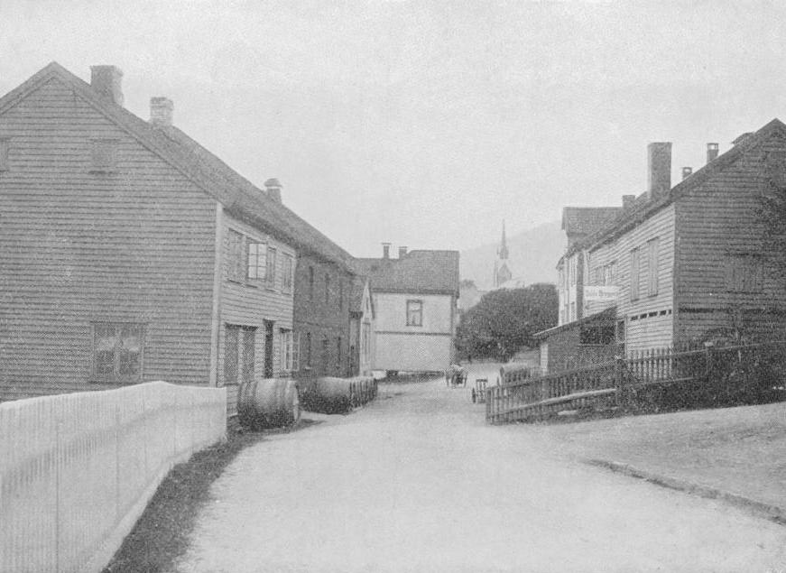 gammelt sort-hvitt bilde som viser et to-etasjes hus med en rekke eiketønner liggende utenfor